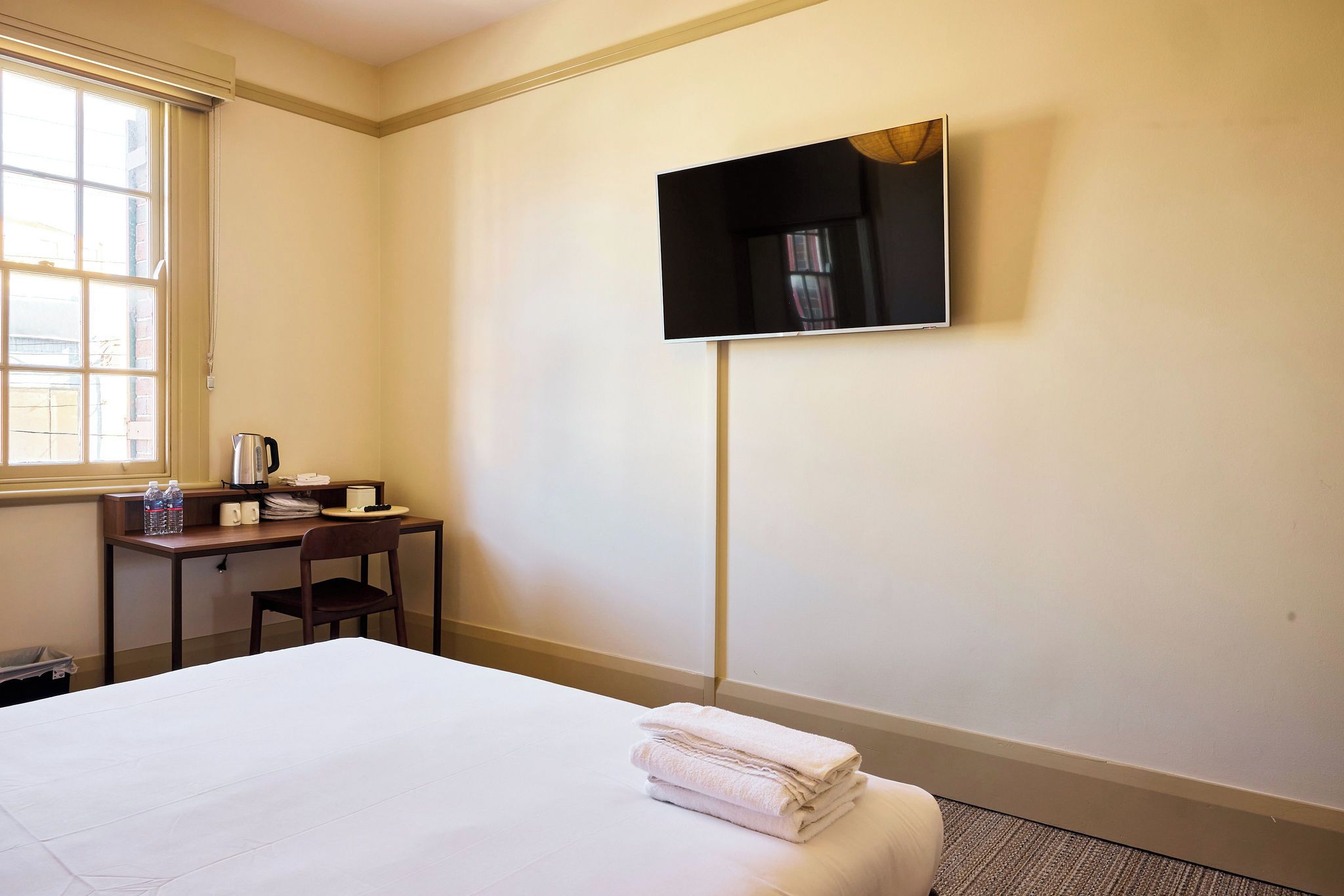 A hotel room with a bed, a desk, a chair, a flat-screen TV mounted on the wall, and a window.