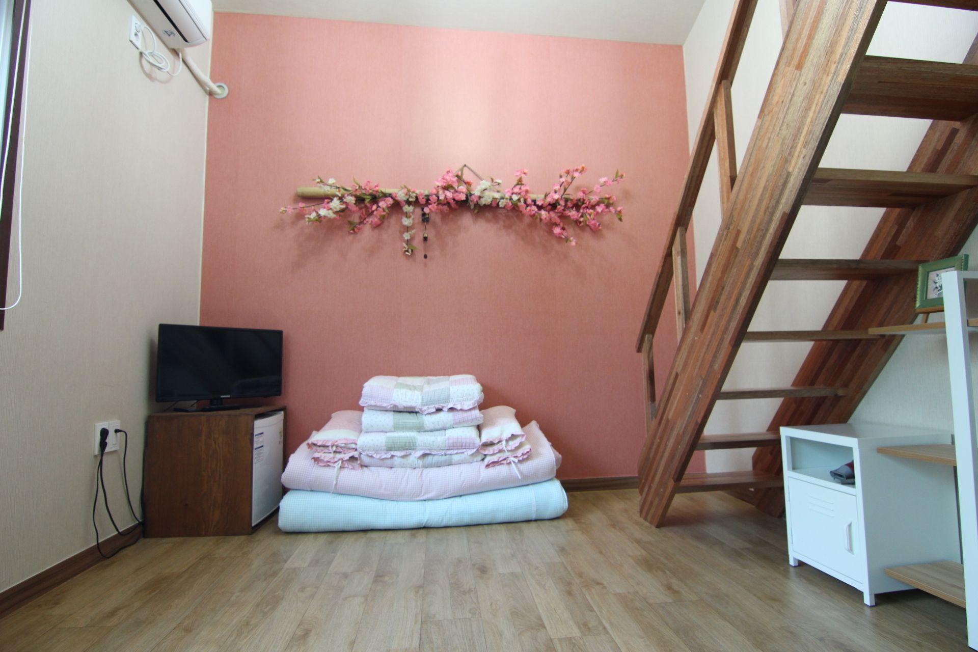 A bedroom with a pink wall, a wooden ladder, a television, and a stack of folded blankets.
