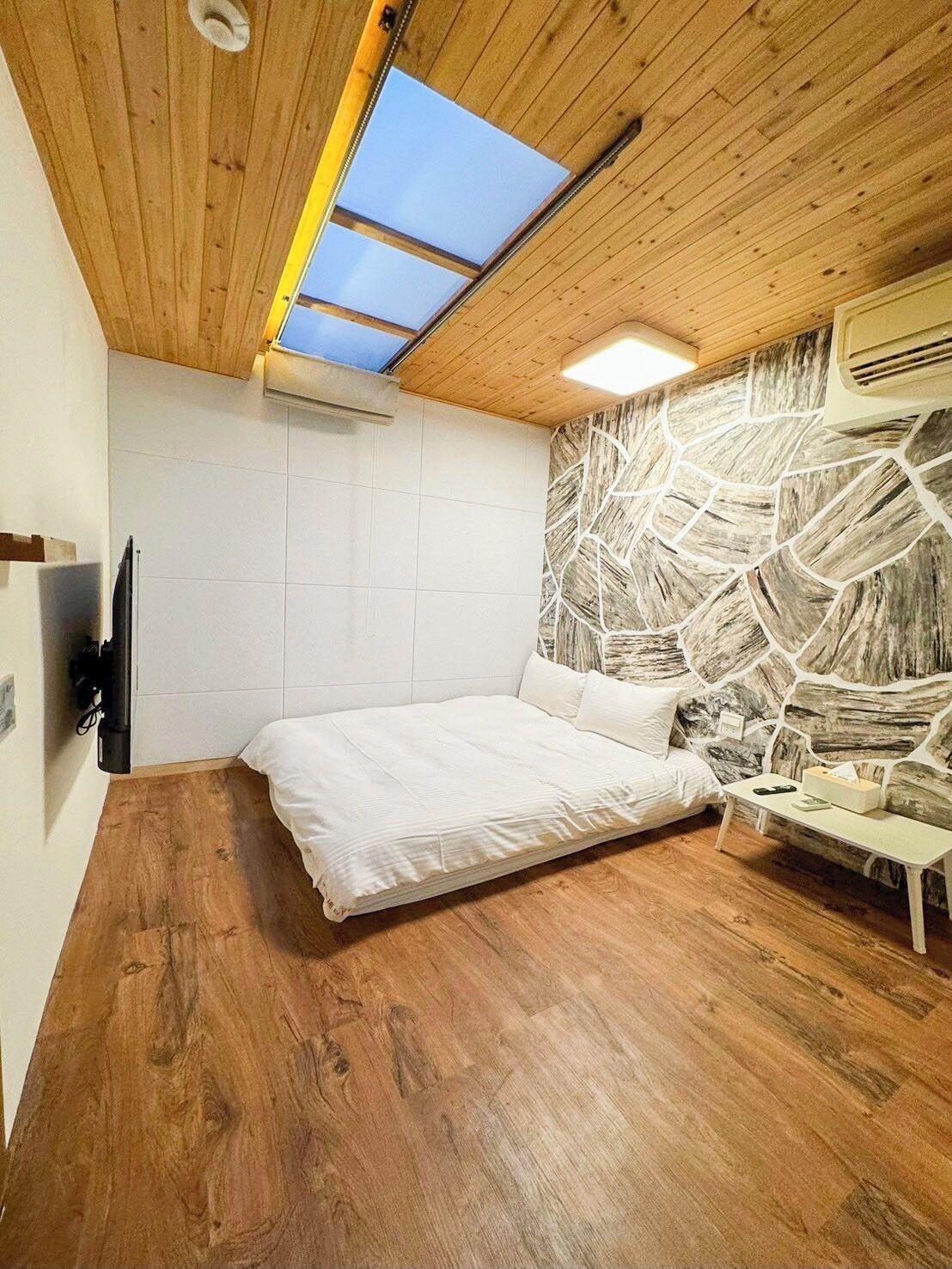 A bedroom with a wooden ceiling, a skylight, a stone-patterned wall, a bed, and a bedside table.