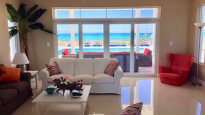 A living room with a white sofa, a red armchair, and a coffee table with decorative items. A large window offers a view of the ocean and a pool.