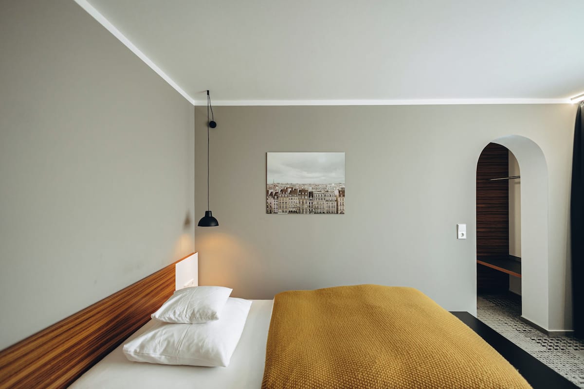 A bedroom with a bed, a headboard, a bedside lamp, a wall-mounted picture, and an arched doorway.