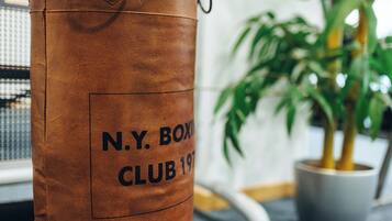 Un saco de boxeo marrón con el texto 'N.Y. BOXING CLUB 197', una planta en maceta y una ventana al fondo.