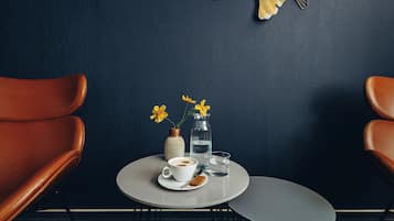 Una pequeña mesa con una taza de café, un vaso de agua, un jarrón con flores y una concha decorativa sobre una pared oscura.