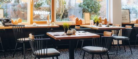 Un restaurante con mesas y sillas, ventanas amplias y lámparas colgantes.