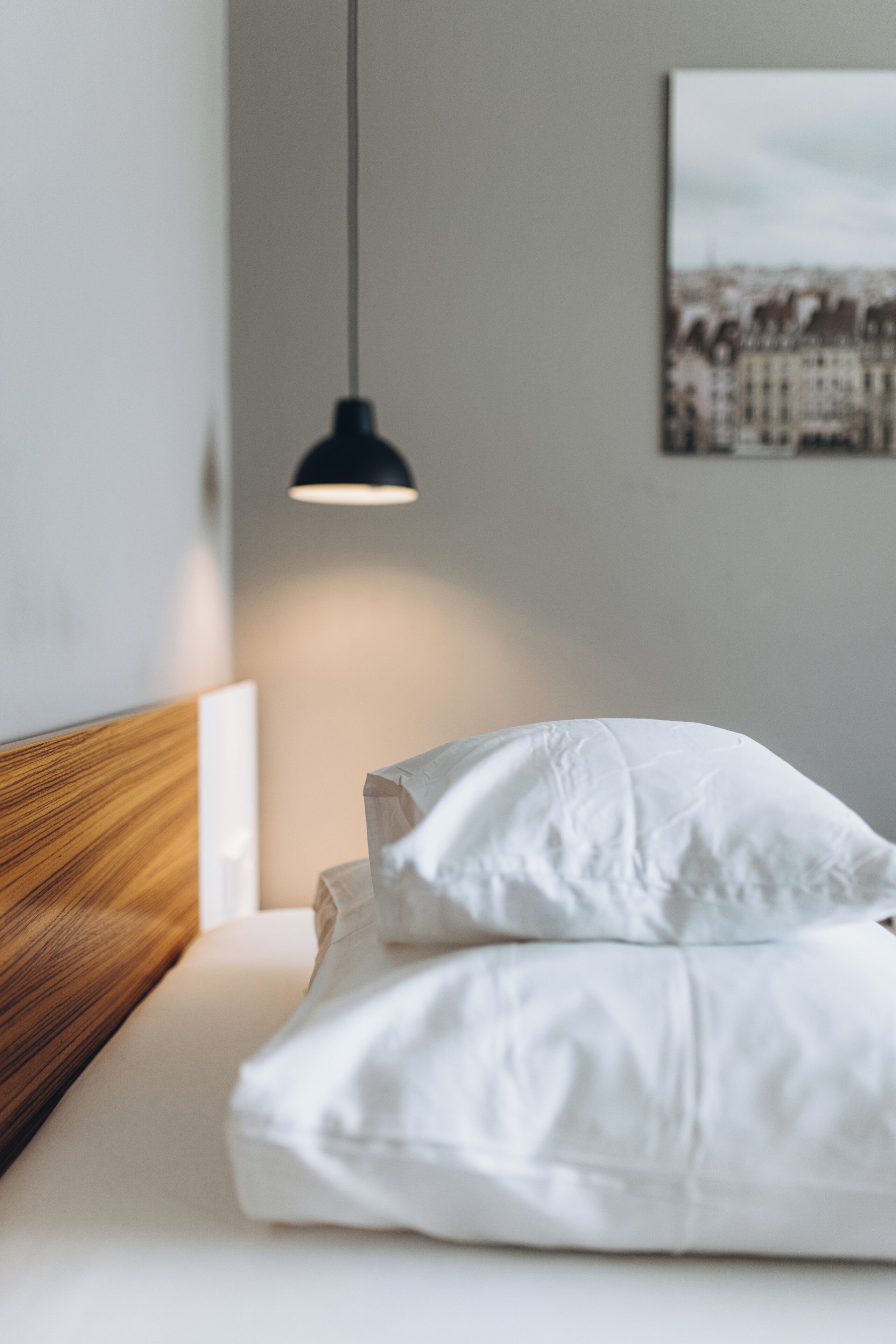 Een net opgemaakt bed met witte lakens, een houten hoofdbord, een hangende吊灯 en een lijst met een afbeelding aan de muur.