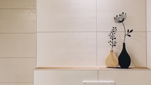 Un baño con pared de azulejos, repisa de madera, un florero negro con planta y un interruptor de luz en la pared.