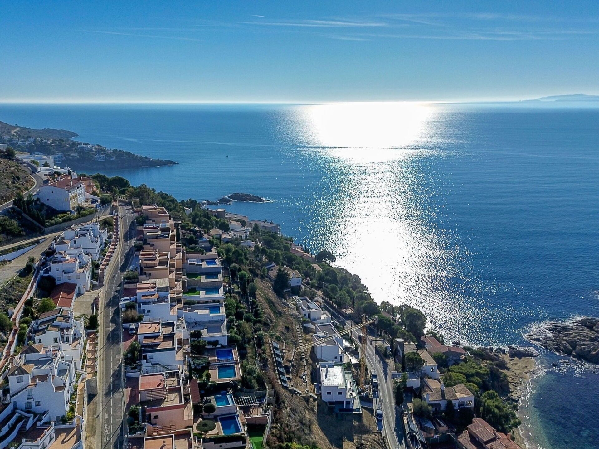 An aerial view of a coastal residential area with houses, a road, and a clear view of the sea.