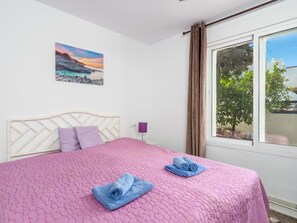 Een slaapkamer met een bed, een schilderij, een raam met gordijnen en uitzicht op groen.