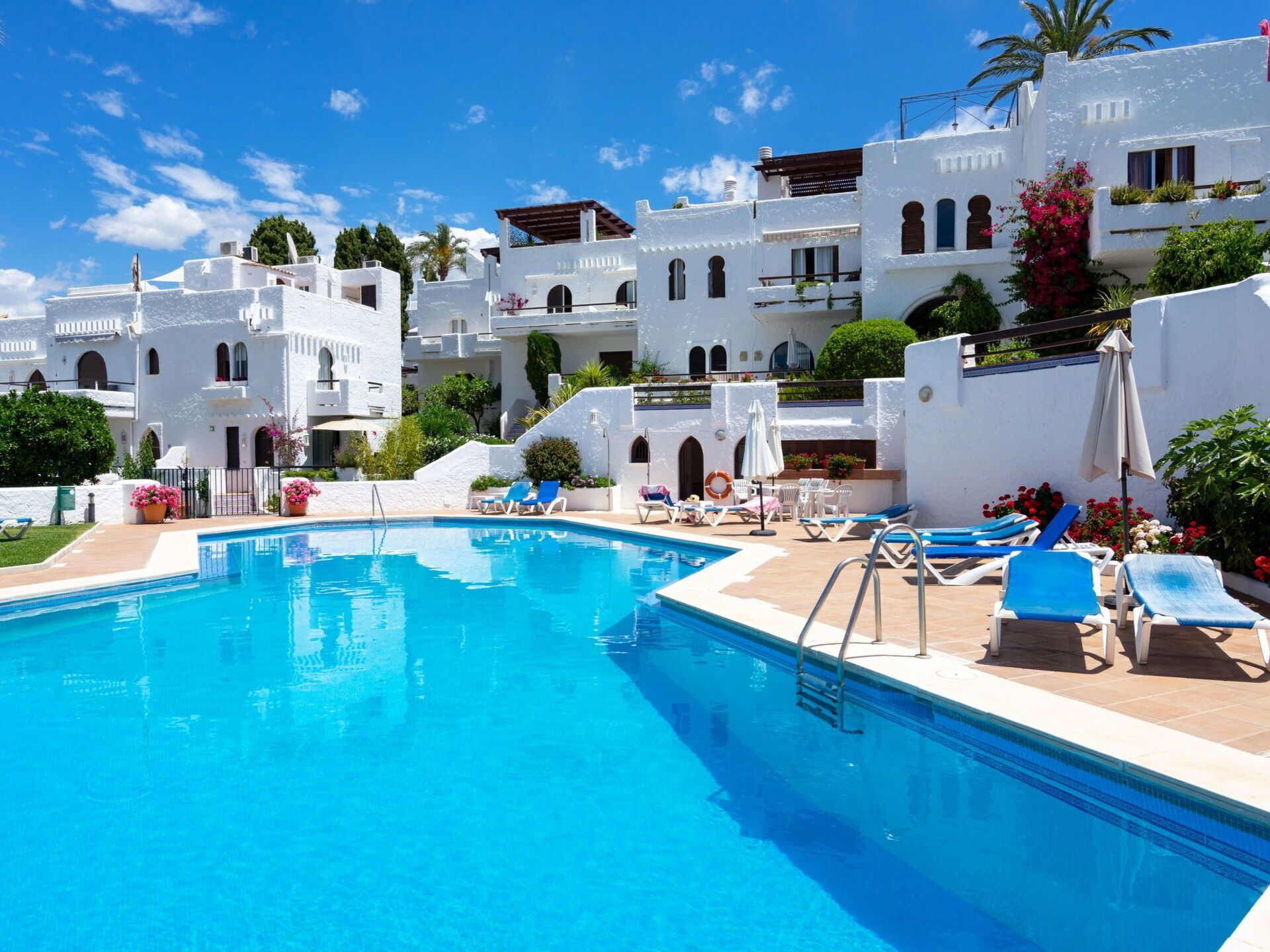 A pool area with white buildings, blue lounge chairs, and a clear blue sky.