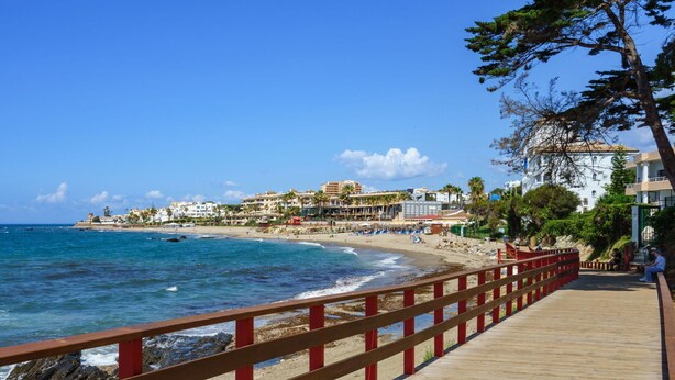 Un muelle de madera con barandas rojas que conduce a una playa, con edificios al fondo.