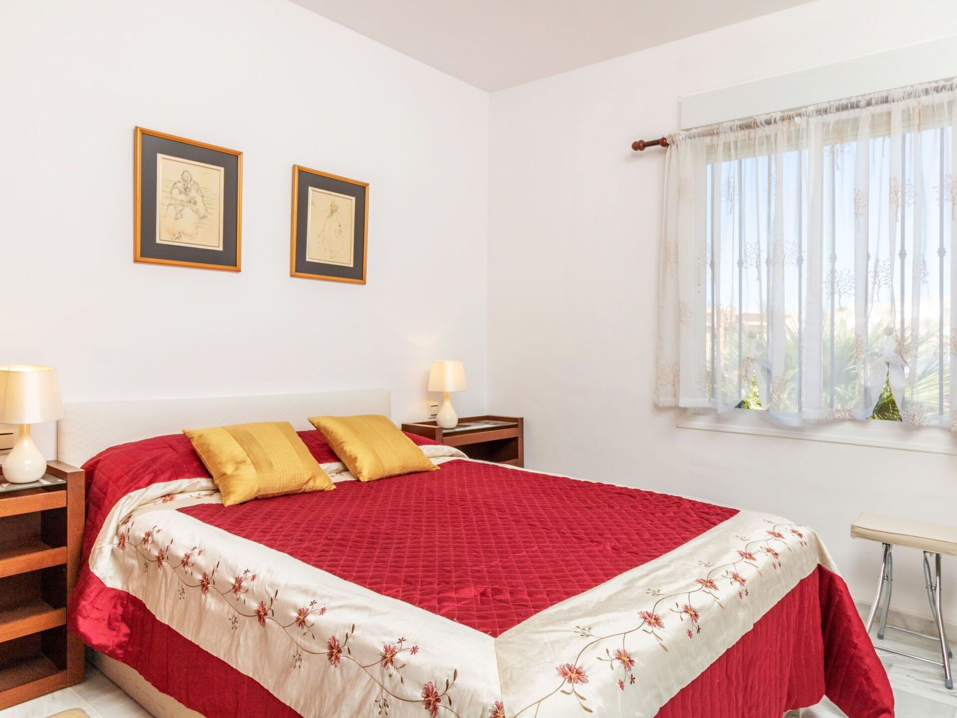 Ein Schlafzimmer mit einem großen Bett, zwei Nachttischen, einem Fenster mit leichtem Vorhang und zwei gerahmten Bildern an der Wand.
