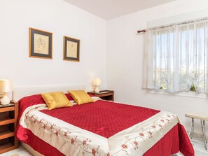 Ein Schlafzimmer mit einem großen Bett, zwei Nachttischen, einem Fenster mit leichtem Vorhang und zwei gerahmten Bildern an der Wand.