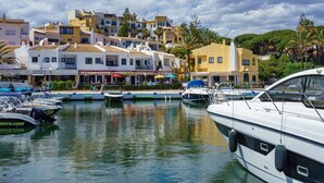 Un puerto deportivo con barcos amarrados, un edificio frente al agua con tiendas, y un cielo despejado.
