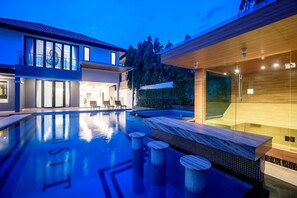 Une maison moderne dotée d’une piscine, d’un espace de spa clos par des parois de verre et d’un coin salon extérieur bien éclairé.