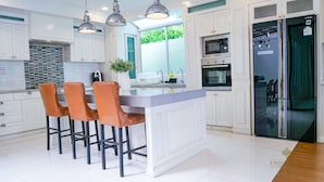 A modern kitchen with white cabinetry, a central island with bar stools, stainless steel appliances, and pendant lighting. - Exquisite Pool Villa C (Pattaya)