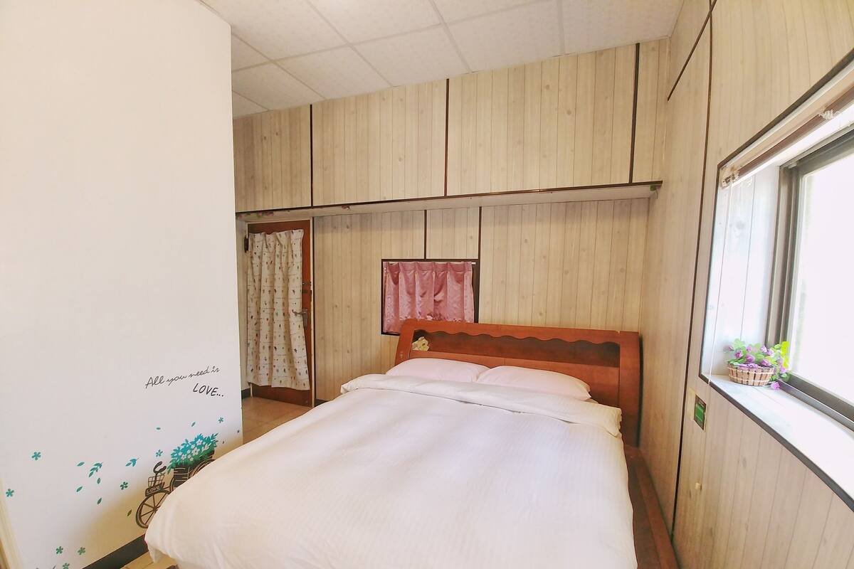 A bedroom with a bed, wooden panel walls, a window with a plant, and a door with a curtain.