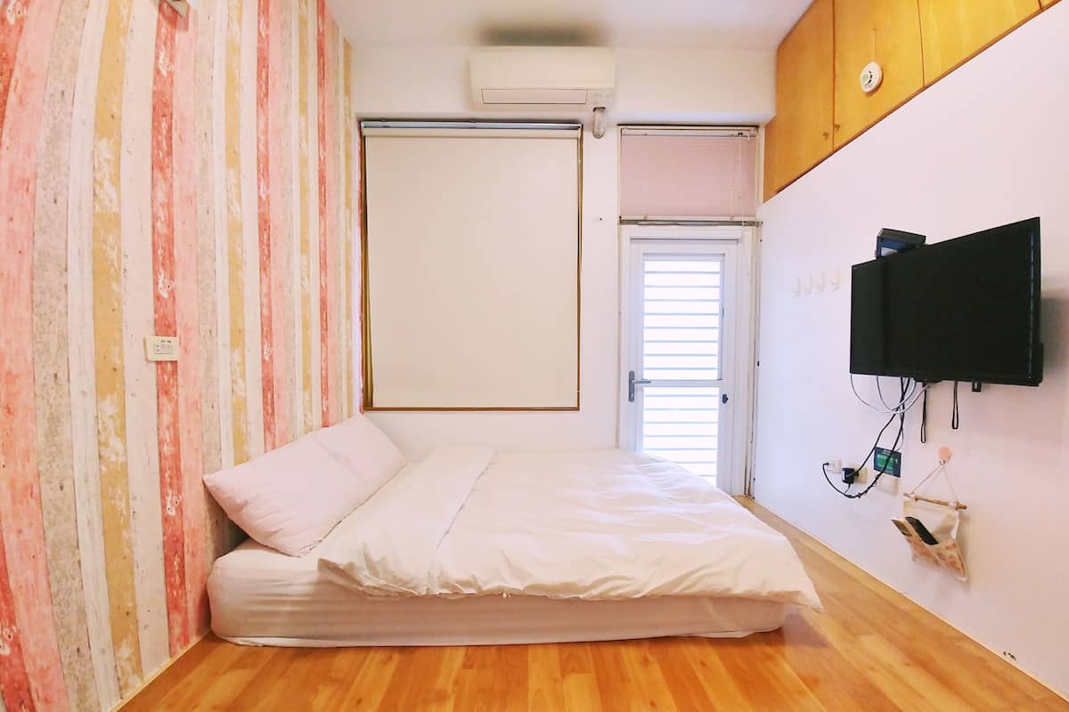 A room with a bed, a wall-mounted TV, a window with blinds, and an air conditioning unit.