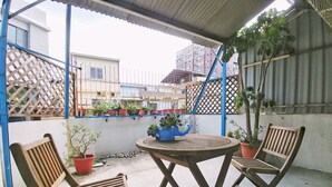 Uma área de estar coberta ao ar livre com duas cadeiras de madeira e uma mesa, cercada por plantas em vasos e uma cerca de metal.