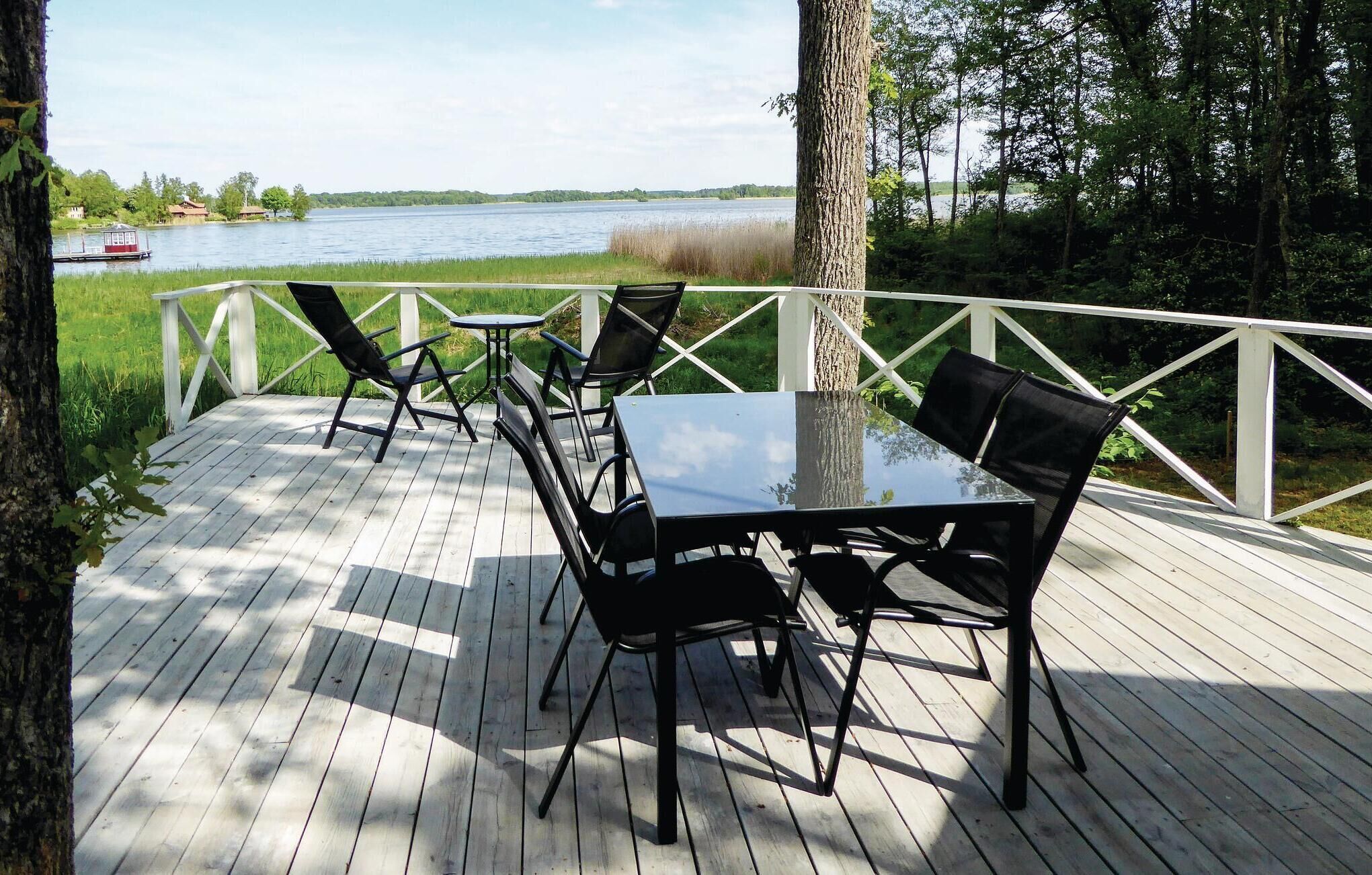 Eine Holzterrasse mit einem Glastisch und schwarzen Stühlen, mit Blick auf einen See, auf dem ein Boot fährt, und Gebäude in der Ferne.