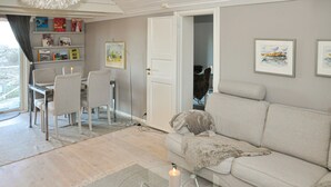 Ein Wohnzimmer mit einer Couch, einem Esstisch mit Stühlen, einem Bücherregal und einer Tür zu einem anderen Raum.