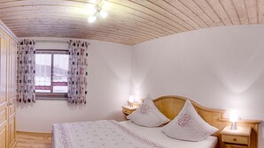 Un dormitorio con techo de madera, una cama con ropa de cama floral, dos mesitas de noche con lámparas y una ventana con cortinas estampadas.