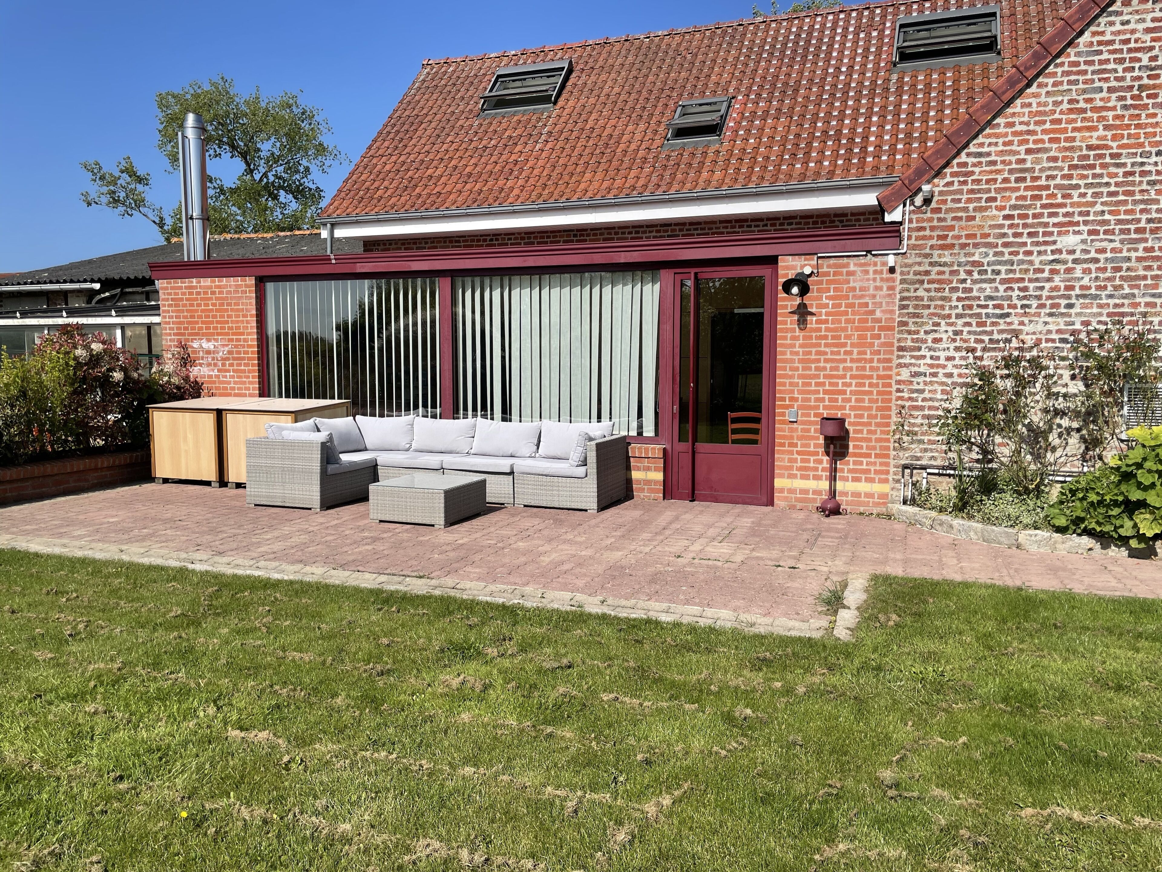 A patio with a sectional sofa and a small table, a brick house with a tiled roof, and a large window with curtains.