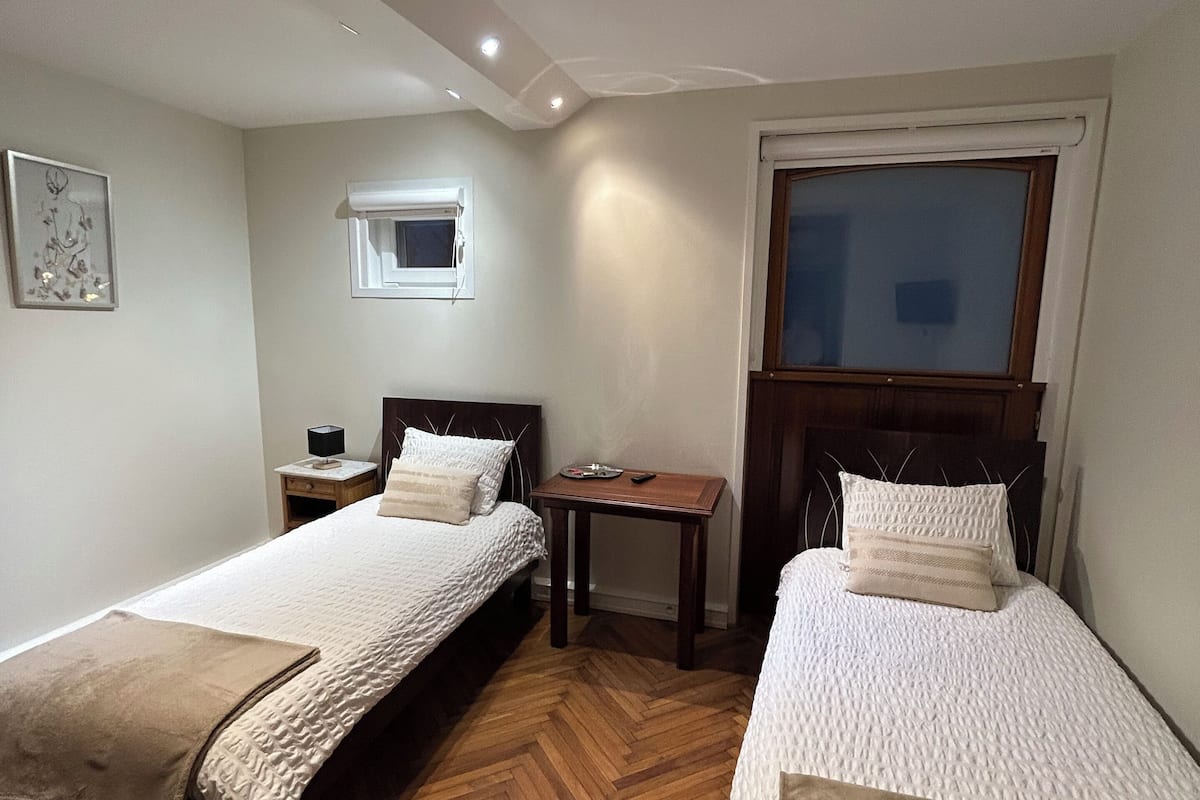 Une chambre d’hôtel avec deux lits simples, une petite table, une table de chevet, une fenêtre et un luminaire au plafond.