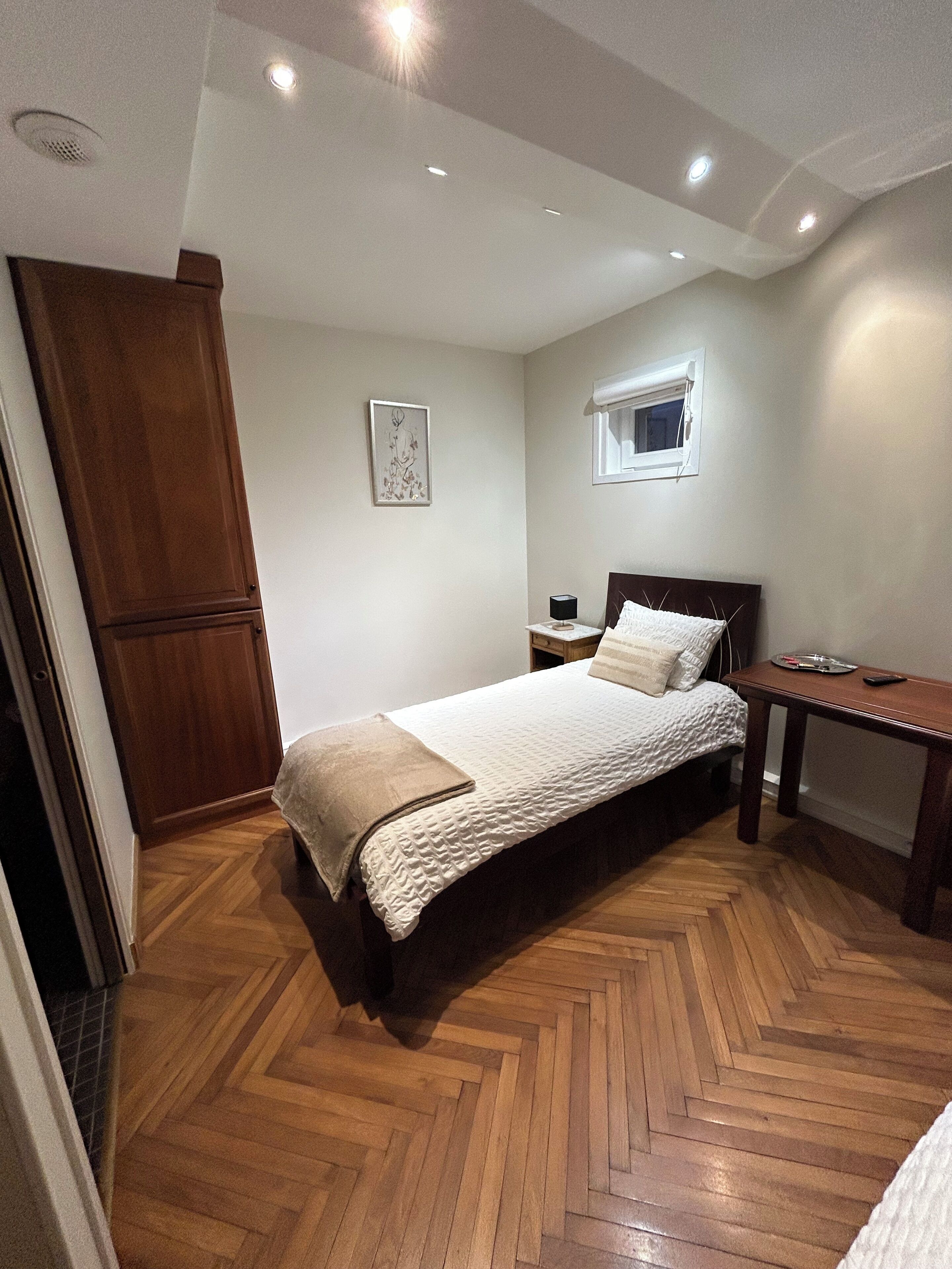 Une chambre avec un parquet en bois disposé en motif «chevrons», un lit recouvert d’une couette blanche, une table de chevet avec une lampe et une armoire en bois.