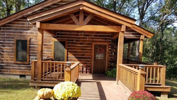 A wooden cabin with a porch, a door with a wreath, and potted plants. - Contemporary Glamping with Native American Appeal (Anna)