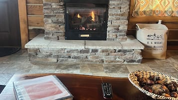 A stone fireplace with a fire, a wooden table with a remote control, a basket of nuts, and a patterned rug.