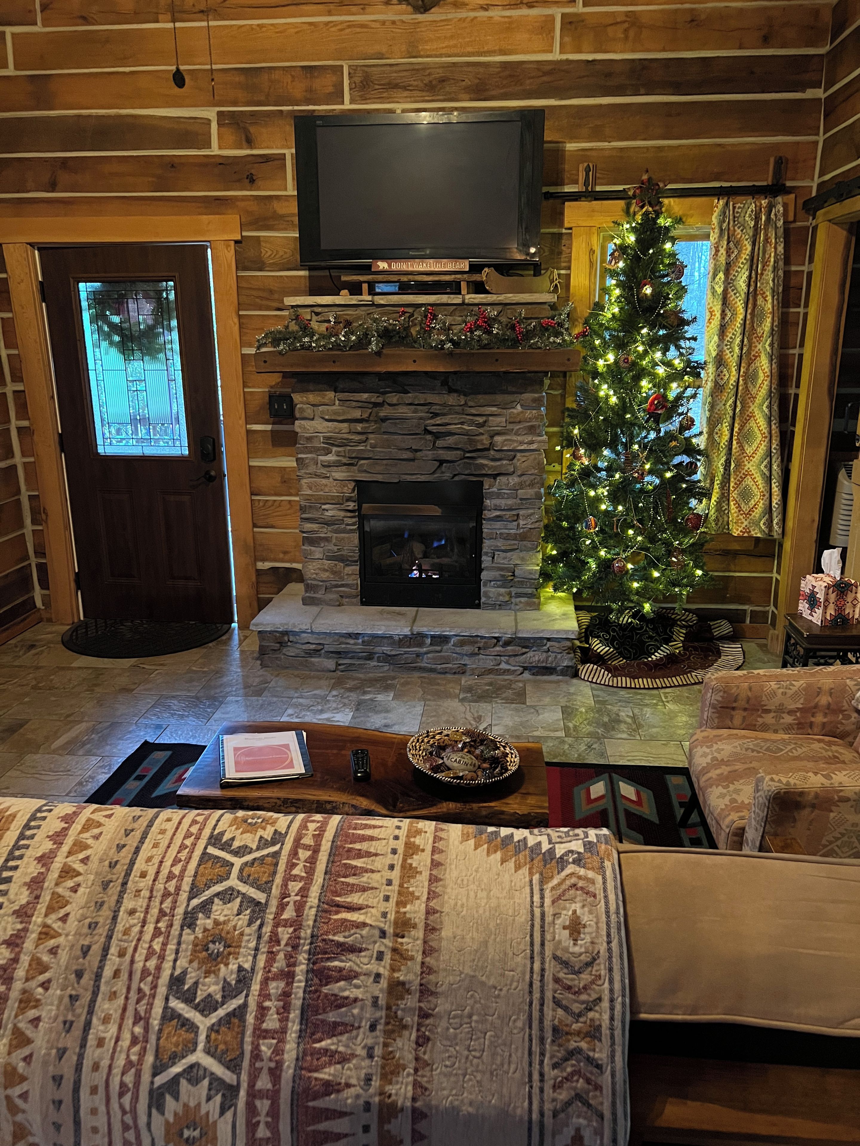 A cozy living room with a stone fireplace, a television mounted above it, and a Christmas tree decorated with lights and ornaments.