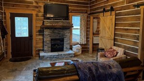 A log cabin interior with a stone fireplace, a mounted television, and a cozy seating area. - Contemporary Glamping with Native American Appeal (Anna)