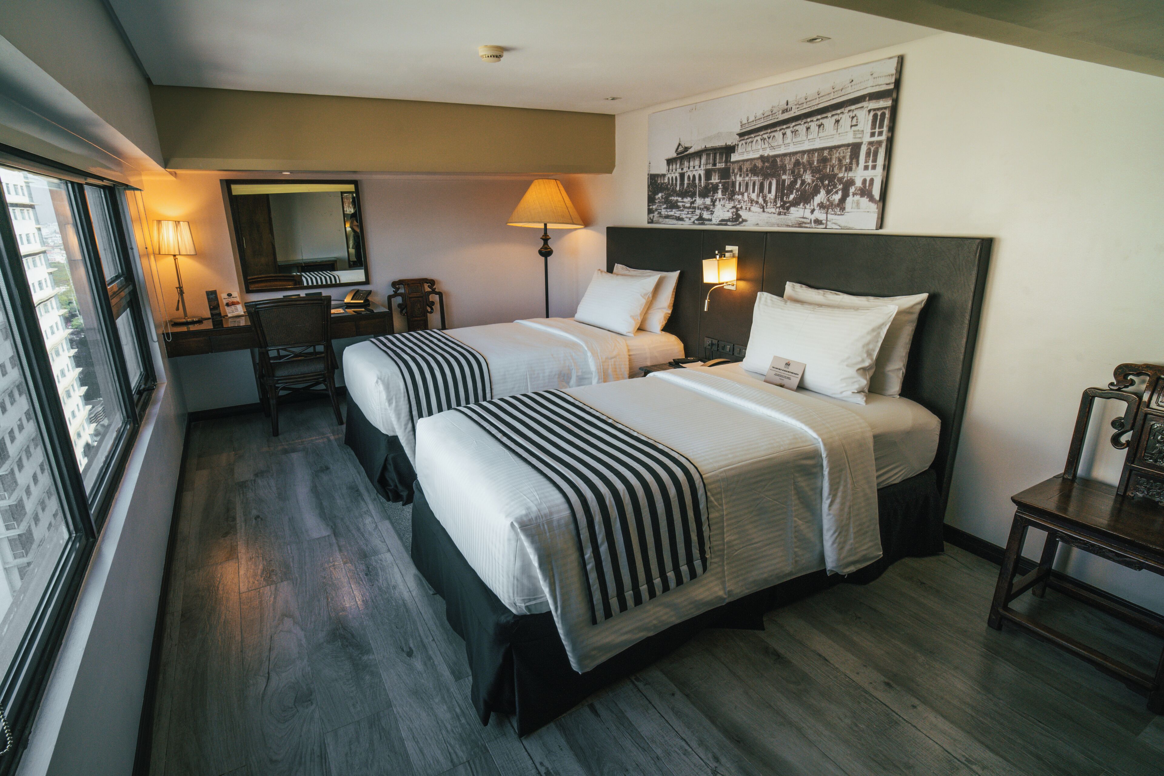 Habitación de hotel con dos camas, un escritorio, una silla, una lámpara y un ventanal grande.