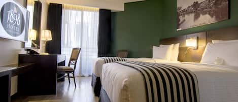 A hotel room with a bed, a desk, a chair, a desk lamp, a wall-mounted TV, and a painting on the wall.