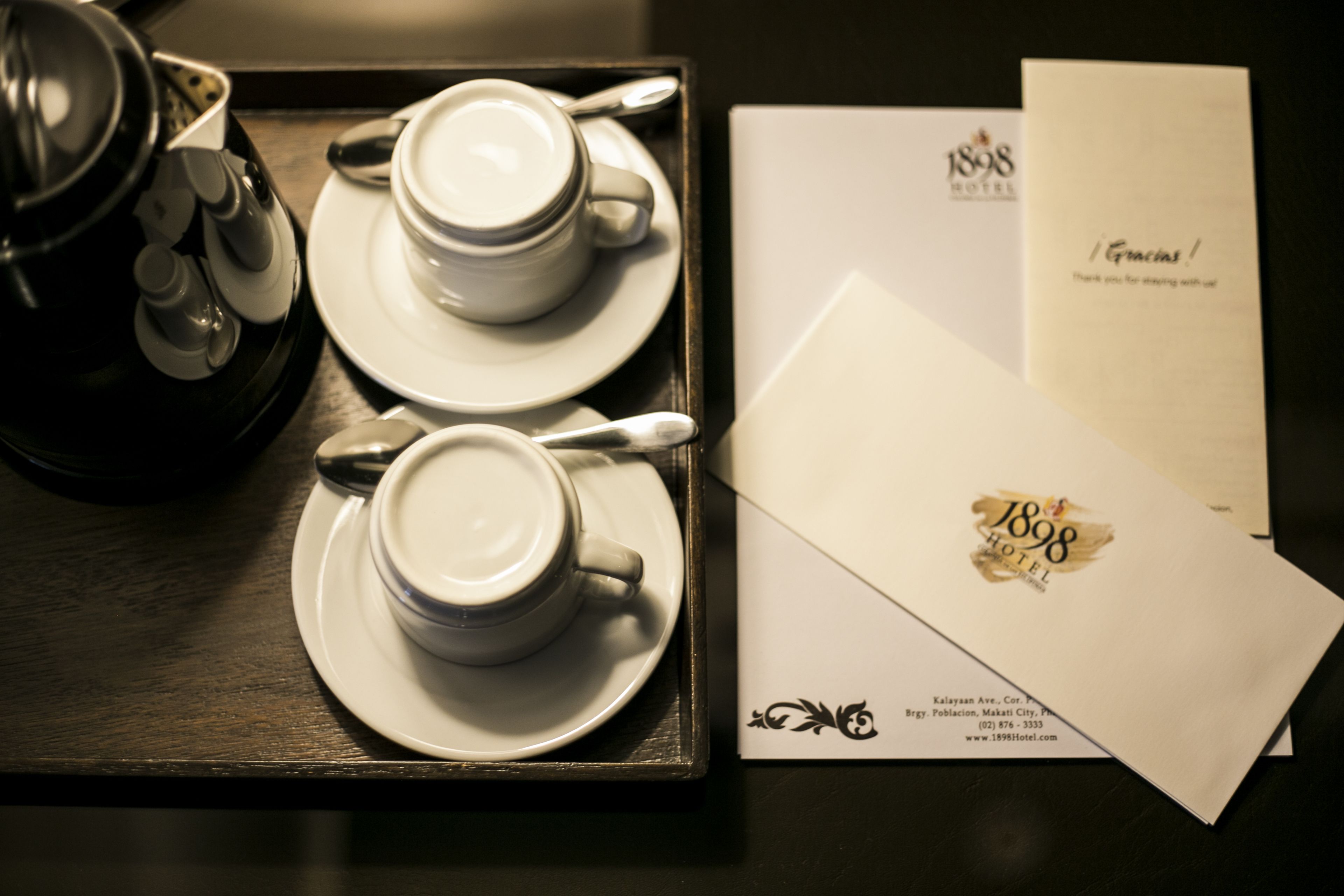 Uma bandeja com duas xícaras de café, um bule e duas chávenas. Também há dois envelopes com o logotipo e o endereço do hotel.