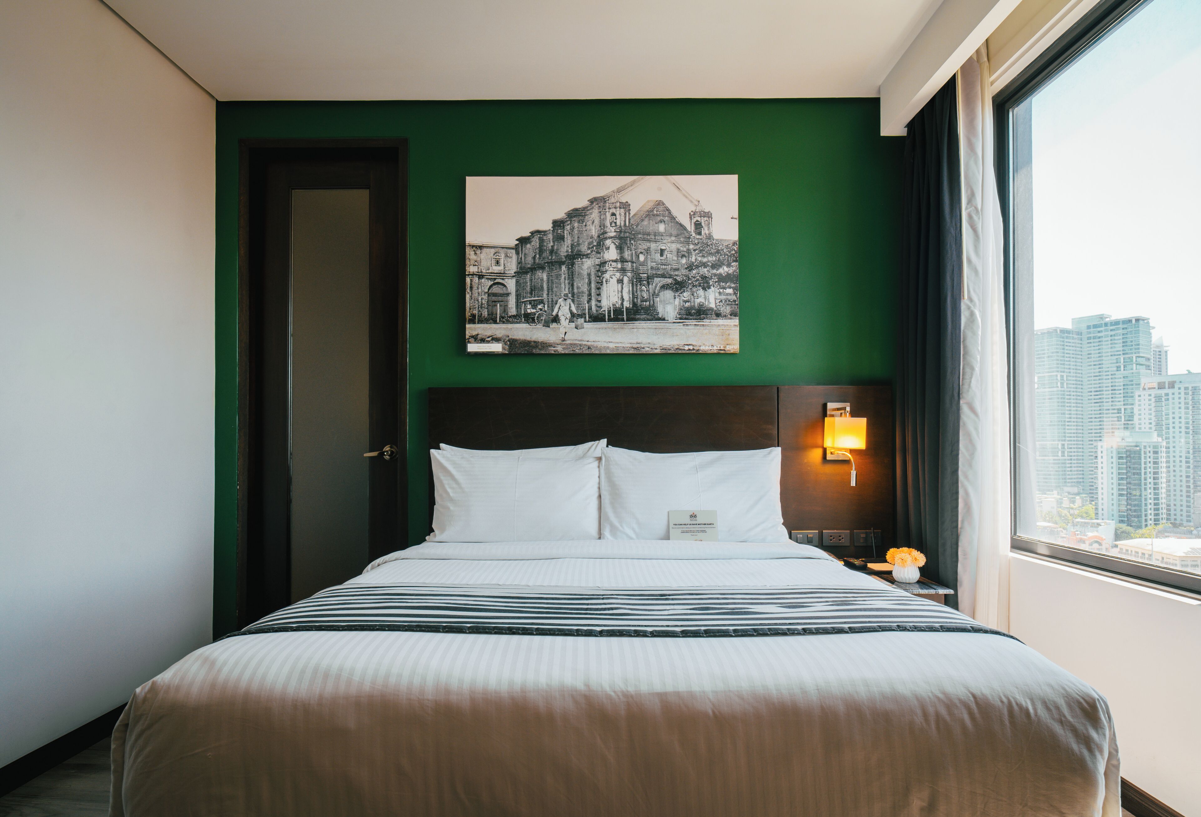 Habitación de hotel con cama, paredes verdes, vista a la ciudad y un cuadro en la pared.