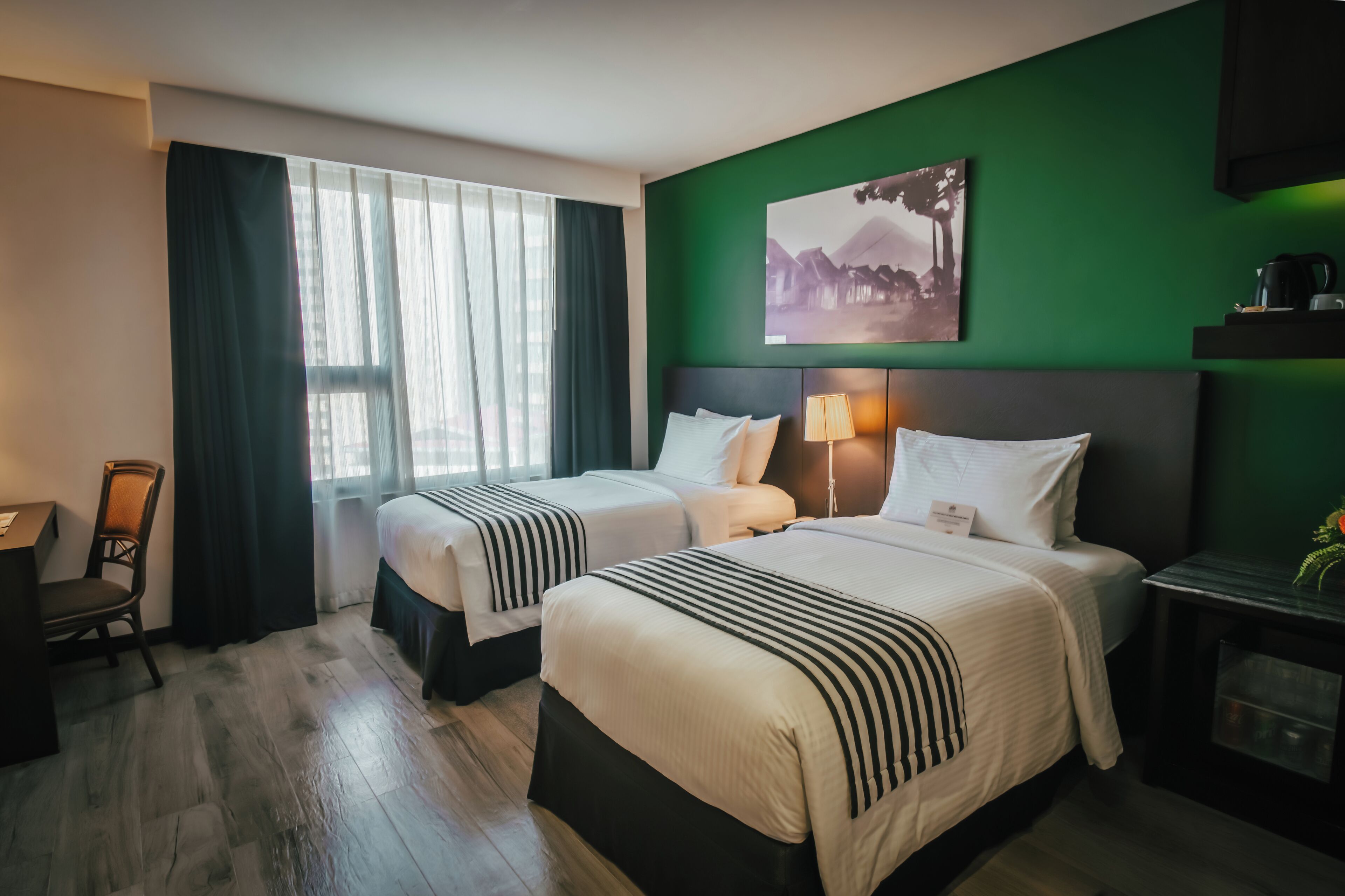 Habitación de hotel con dos camas, un escritorio, una silla, una pared de acento verde y un cuadro.
