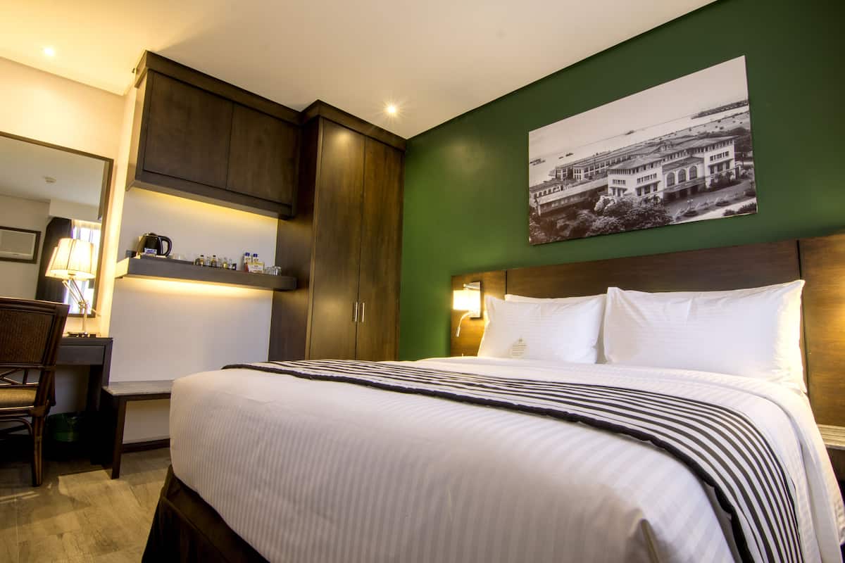 Une chambre d’hôtel avec un grand lit, un bureau avec une chaise, une table de chevet avec une lampe et un mur vert orné d’un tableau encadré.
