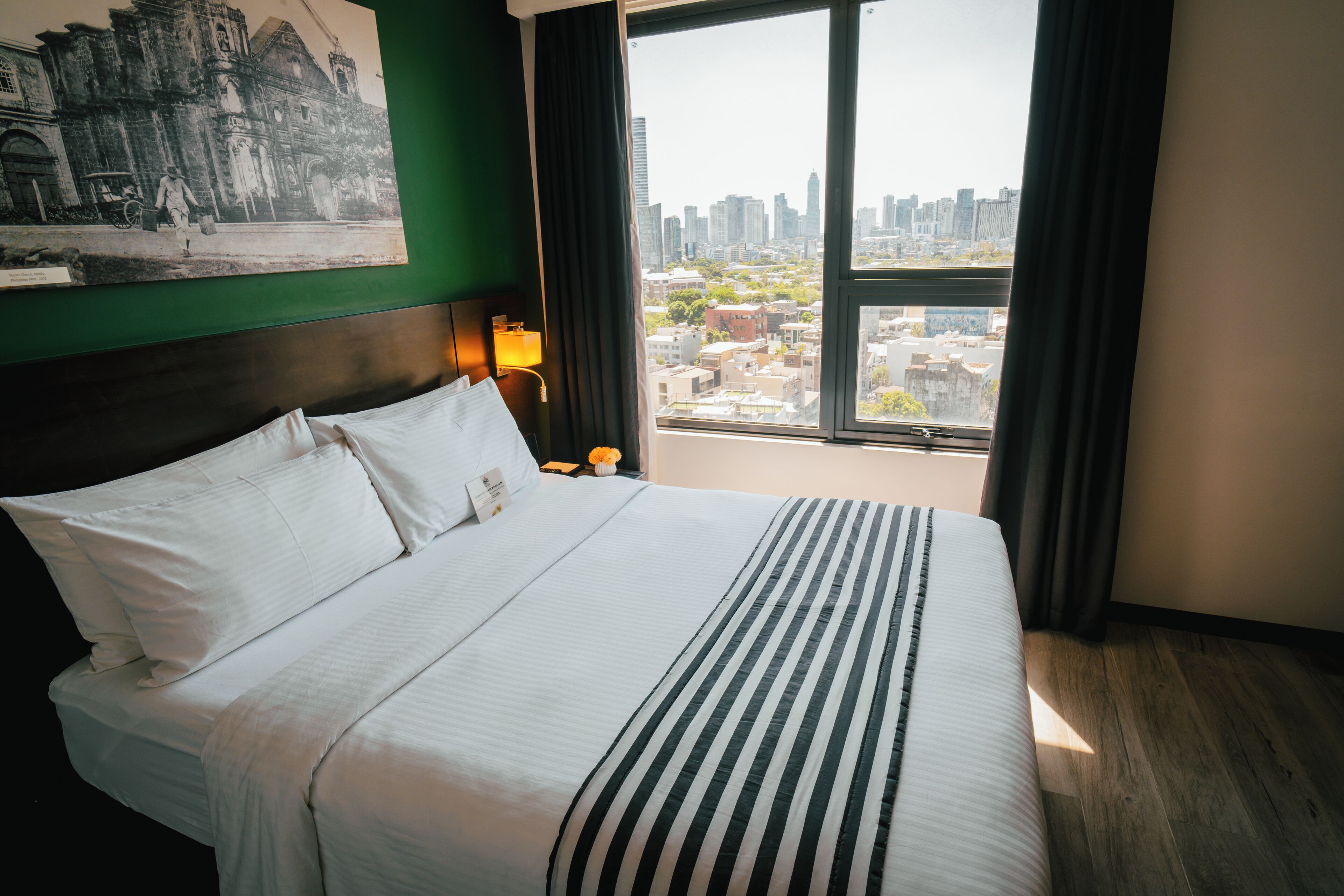 Habitación de hotel con una cama grande, vistas de la ciudad, una fotografía histórica enmarcada en la pared y una mesita de noche con lámpara.