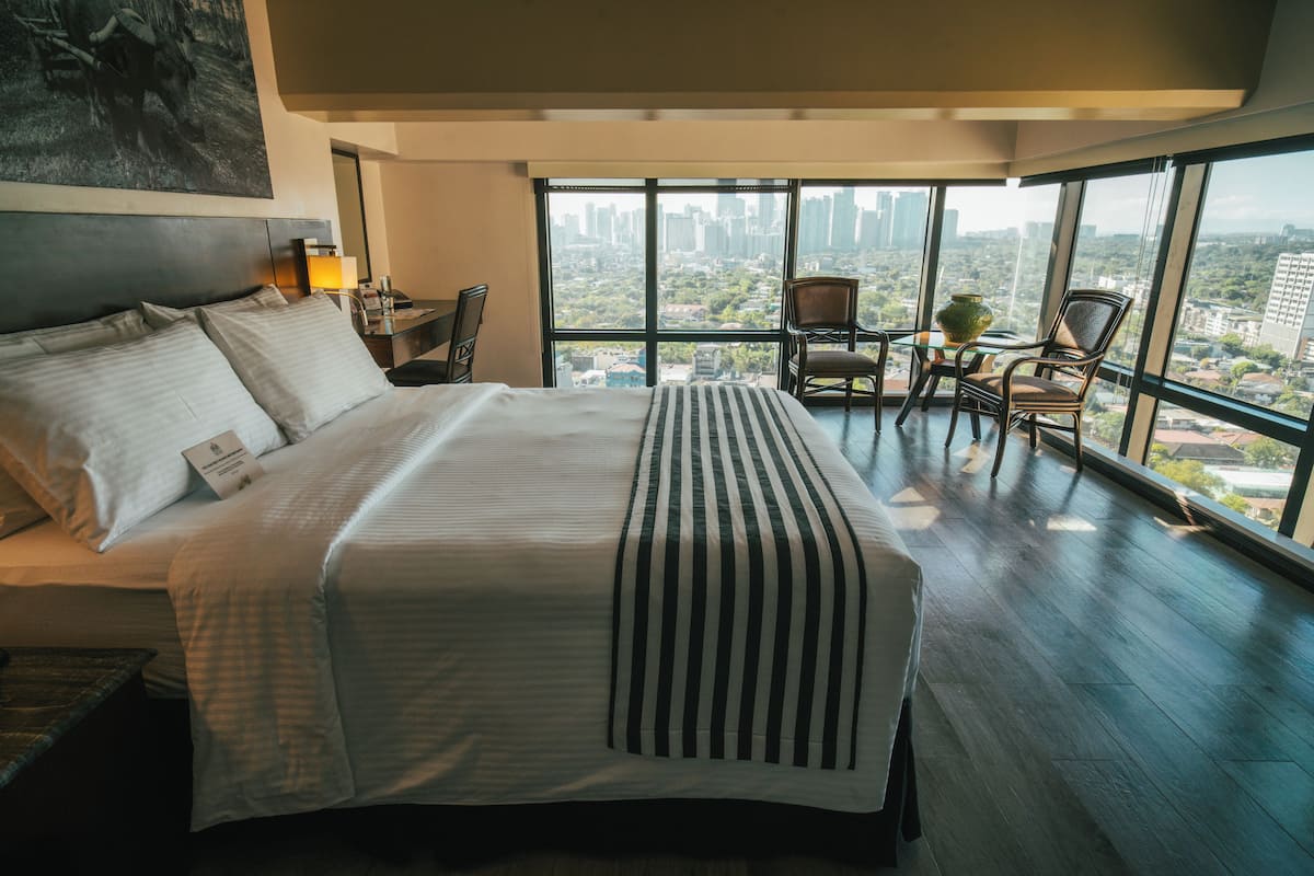 Une chambre d’hôtel moderne avec un grand lit, un bureau avec une chaise, une grande fenêtre donnant sur la ville et une petite table avec un vase.
