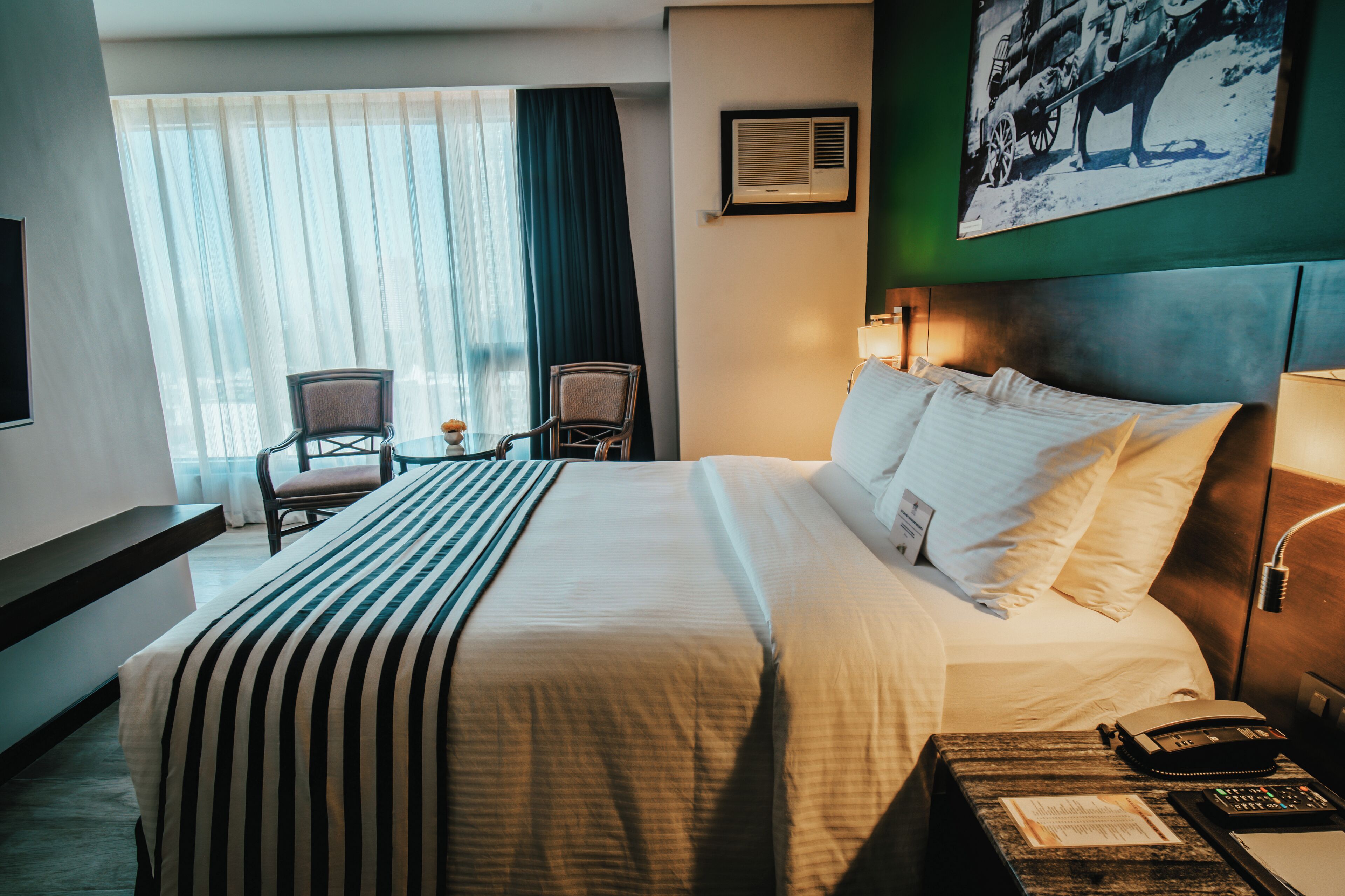 Een hotelkamer met een groot bed, twee stoelen, een bureau met telefoon en een wandairconditioner.