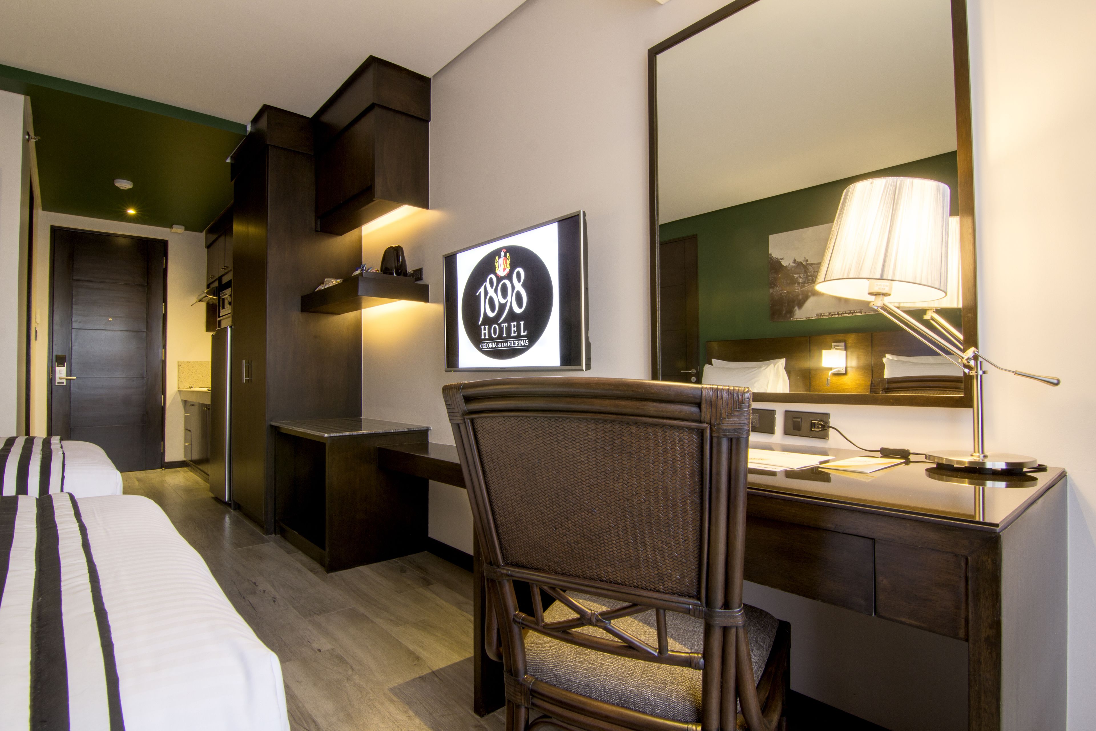 Habitación de hotel con cama, escritorio, silla, espejo y cartel con el nombre del hotel.