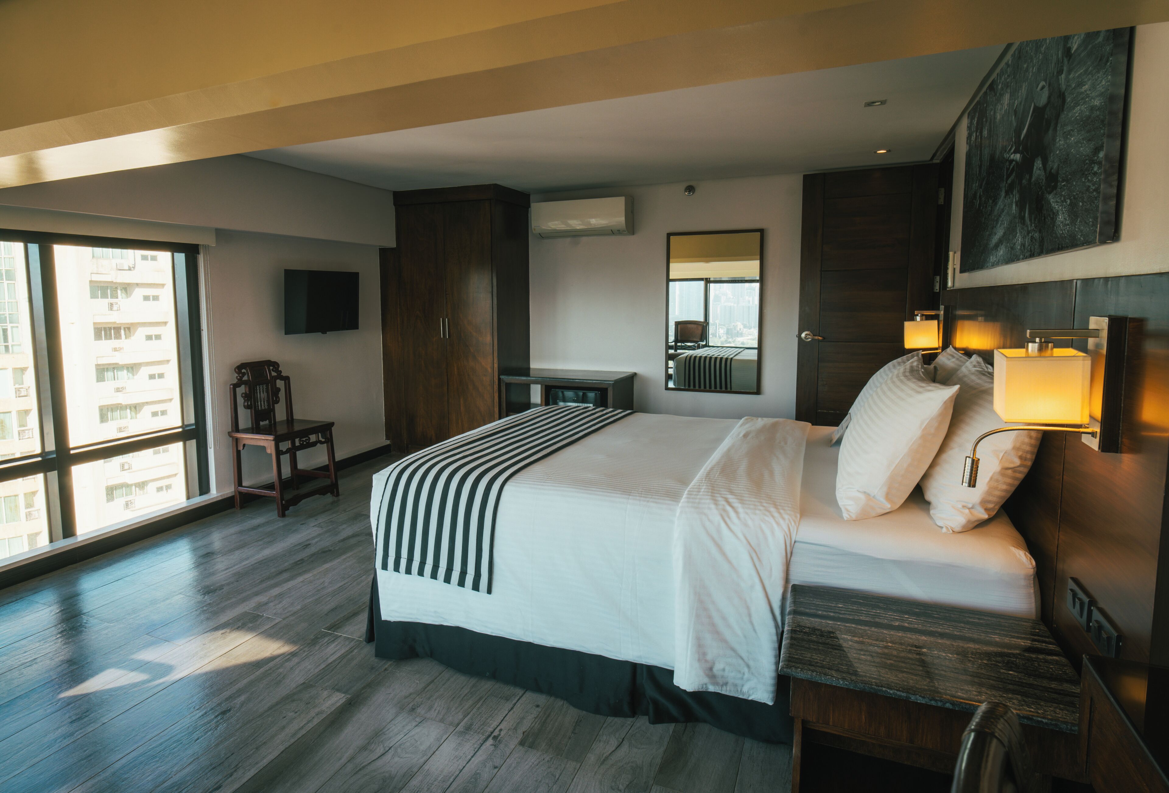Habitación de hotel moderna con una cama grande, un escritorio, una silla, un televisor y un amplio ventanal con vistas a la ciudad.