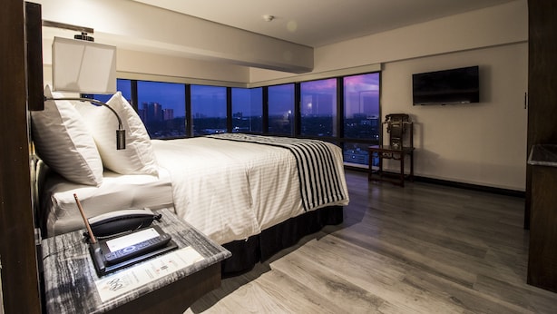 Een moderne hotelkamer met een groot bed, een bureau met telefoon en notitieboekje, een flatscreen televisie en stadszicht door grote ramen.