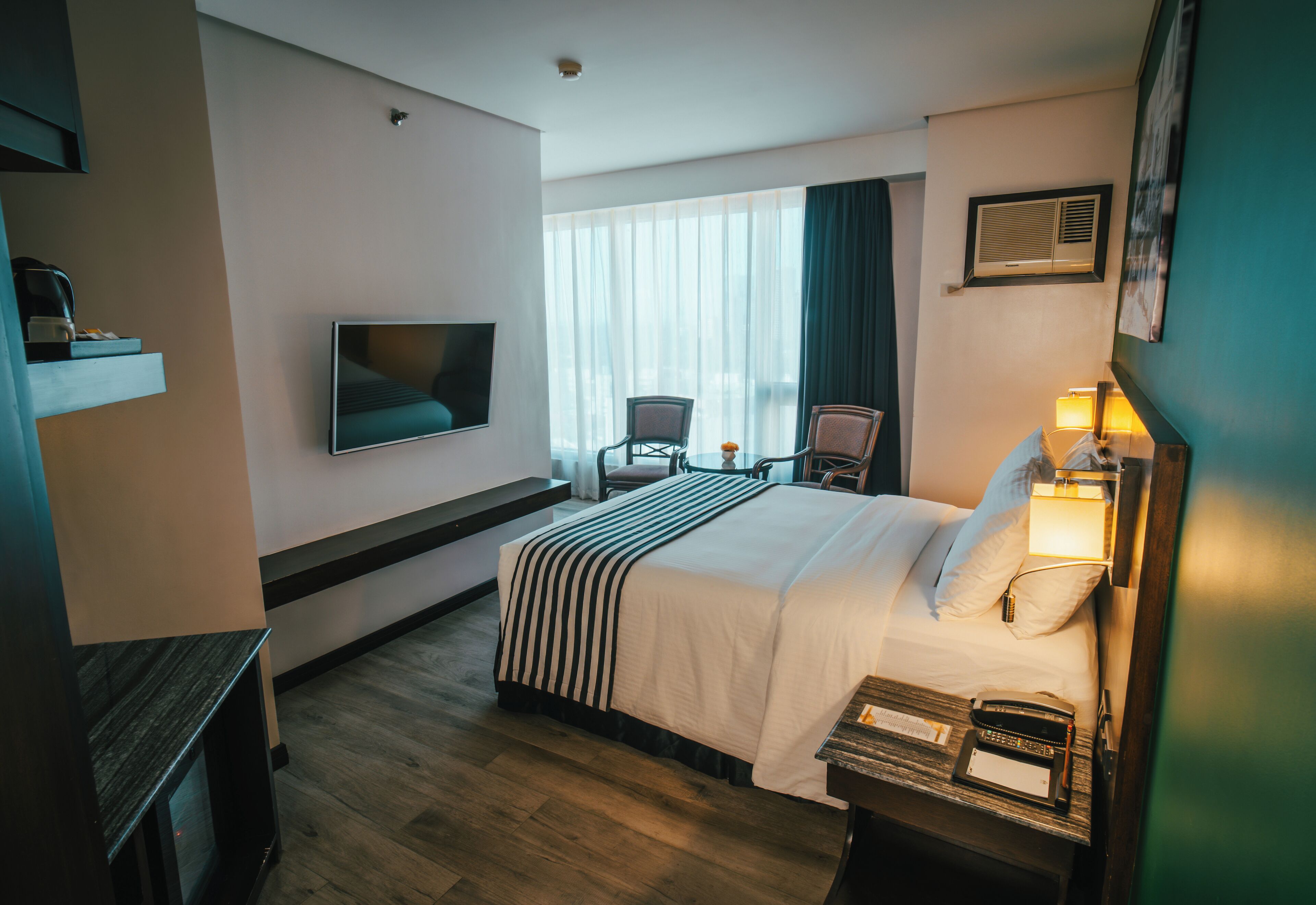 A hotel room with a bed, a TV mounted on the wall, a desk with a lamp, and a chair.