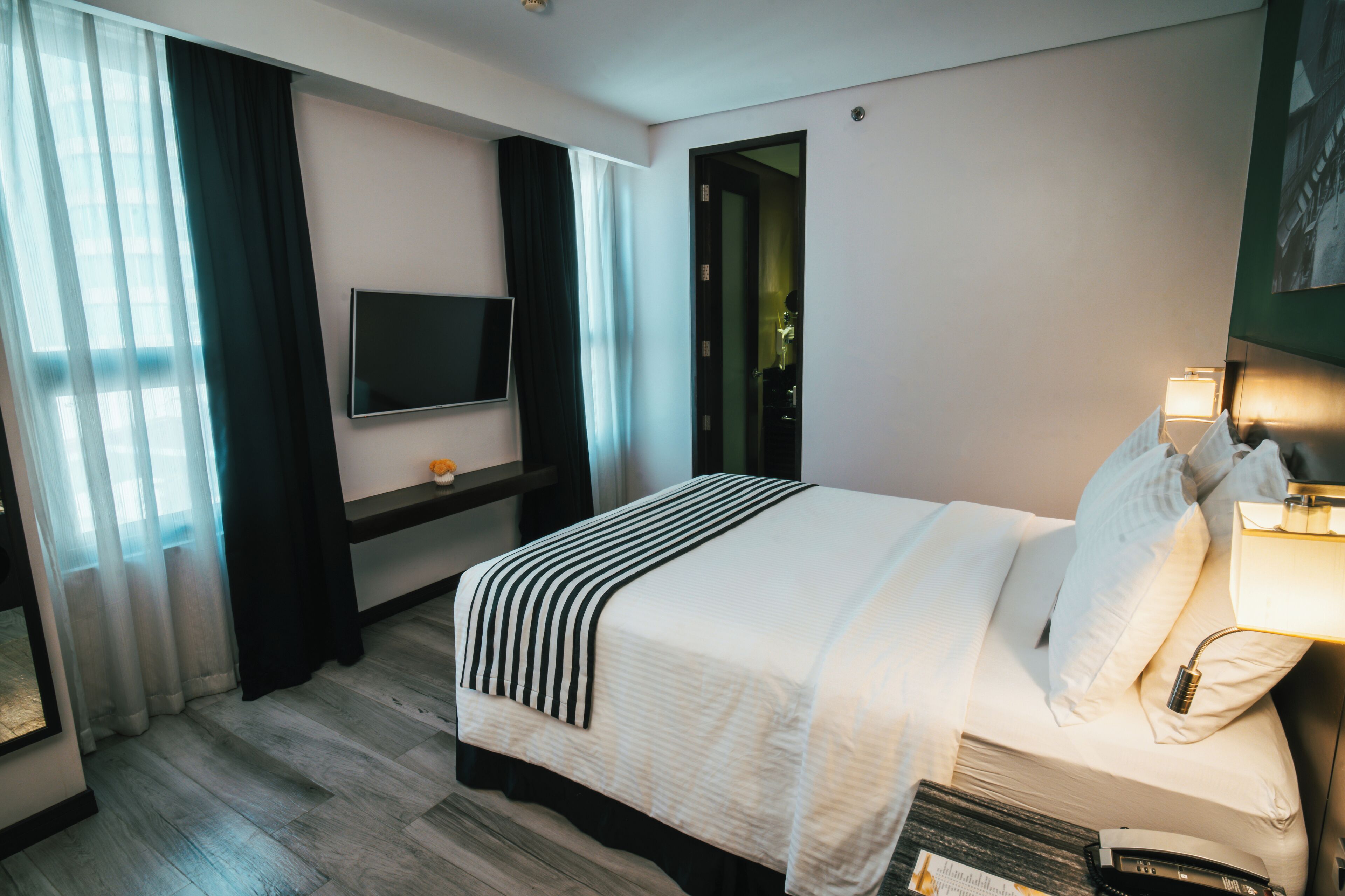 Habitación de hotel con una cama grande, un televisor de pantalla plana montado en la pared, una mesita de noche con lámpara y una puerta que da a otra habitación.