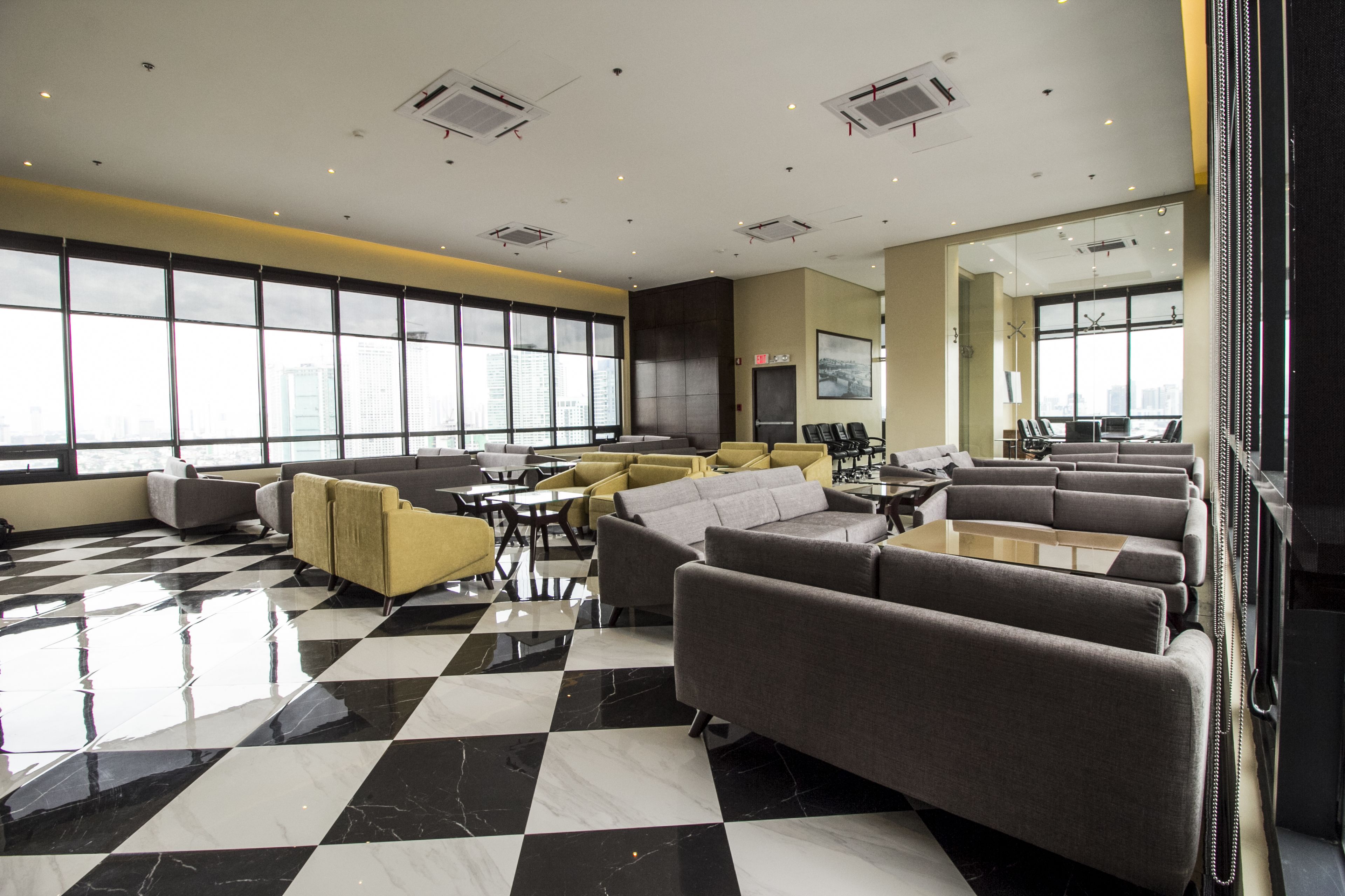 A spacious lounge area with checkered flooring, multiple seating arrangements, and large windows offering city views.