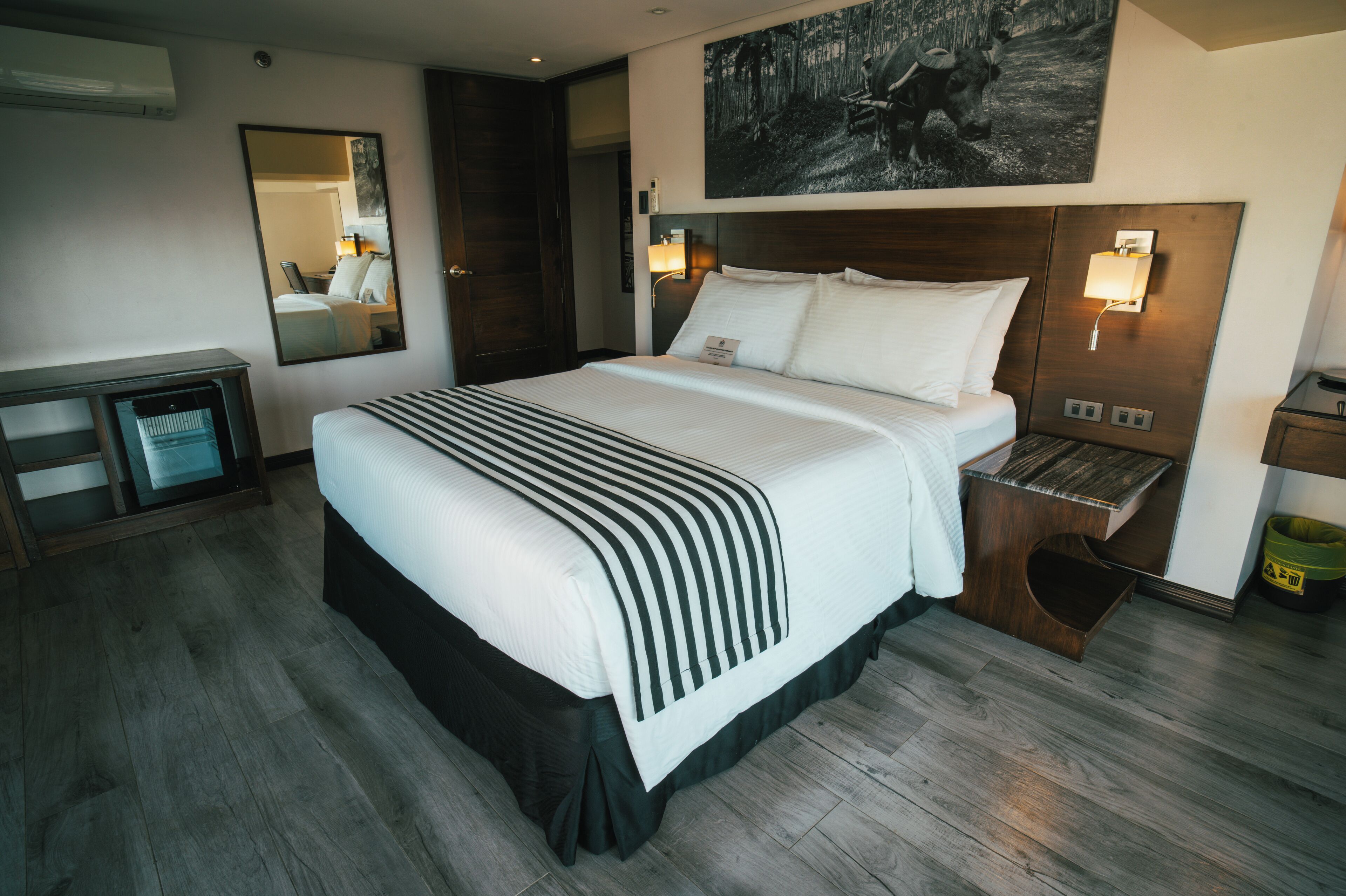 A hotel room with a large bed, a wooden desk, a mirror, a nightstand with a lamp, and a painting on the wall.