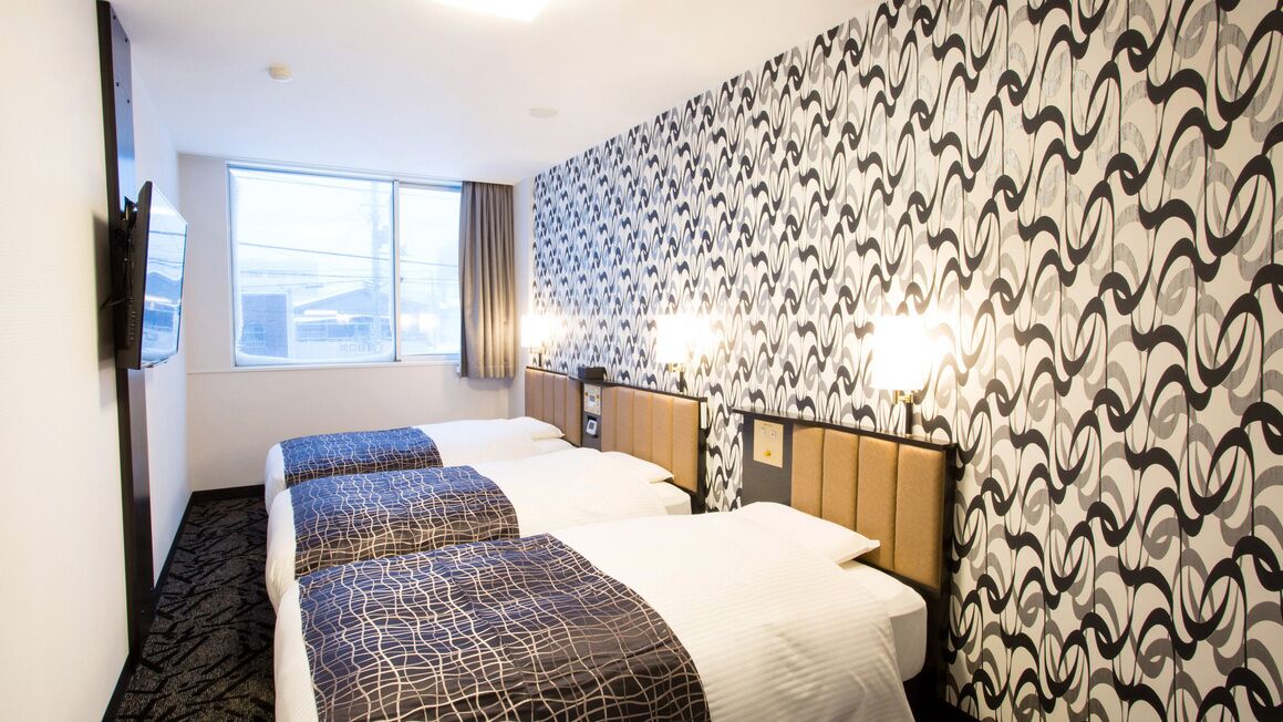 Une chambre d’hôtel avec trois lits, une grande fenêtre, un papier peint à motifs et deux lampes de chevet.