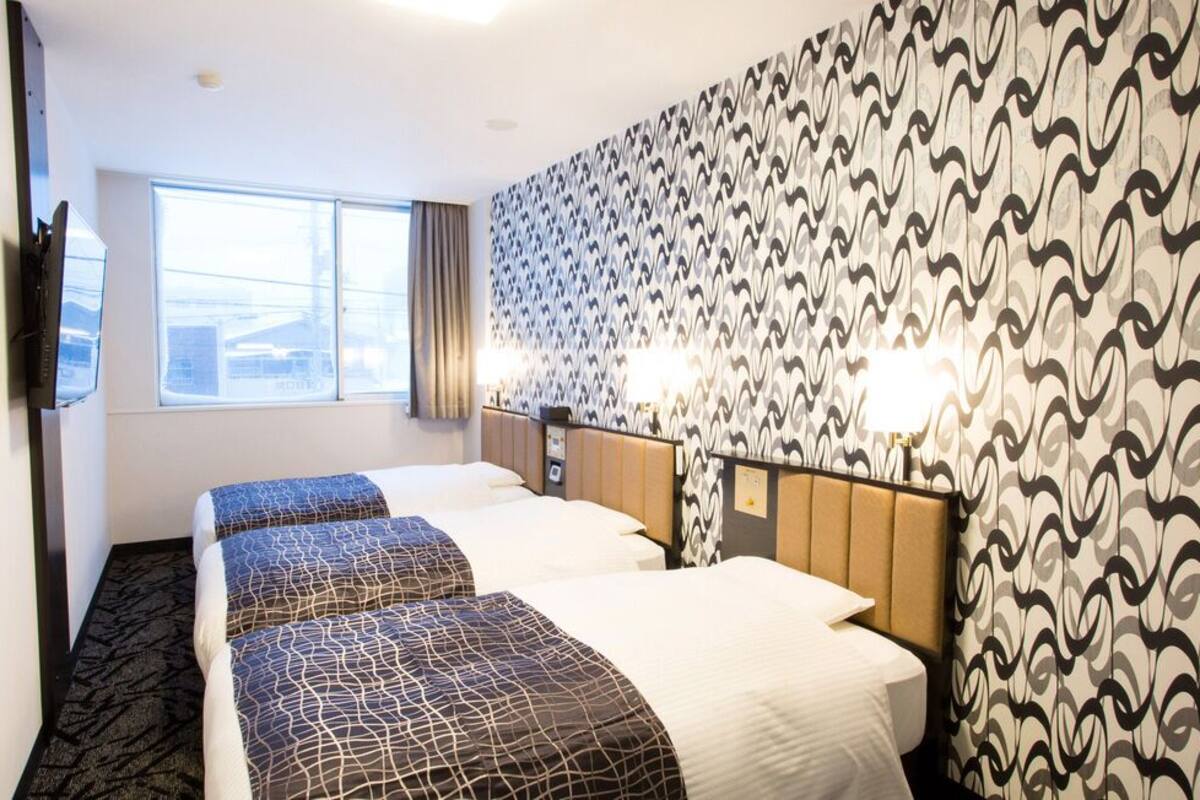 Une chambre d’hôtel avec trois lits, une grande fenêtre, un papier peint à motifs et deux lampes de chevet.